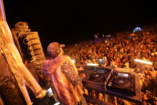 A DJ performs for a large crowd at night during Burning Man 2017 in Lovelock, Nevada. The DJ is wearing a fur vest and headphones, with two