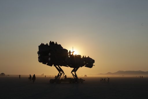A large, mechanical, dinosaur-like art car, carrying passengers, is silhouetted against the setting sun on the playa at Black Rock City, Nev