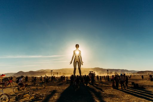 The sun sets behind the iconic "The Man" sculpture at Burning Man 2015, casting long shadows across the playa as a crowd gathers and cyclist