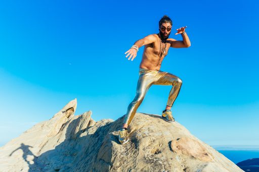 A shirtless man with a beard and bun, wearing shiny gold leggings and sneakers, poses dynamically on a rocky outcrop against a clear blue sk