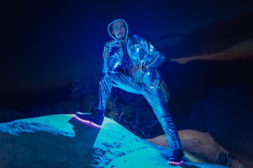 A man in a shiny silver outfit with glowing shoes poses on a rock at night.