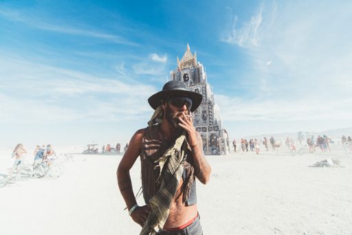 A man smokes a cigarette in front of the "Temple" structure at Burning Man 2015. The vast playa stretches out behind him, dotted with people