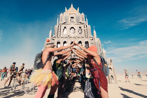 A group of people form a circle with their hands touching overhead in front of a large, ornate structure at Burning Man in 2015. The festiva