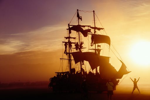 A silhouette of a pirate ship art installation with people climbing its rigging is set against a vibrant sunset at Burning Man. The sun cast