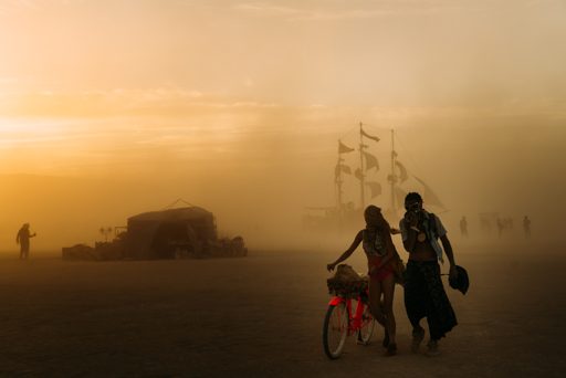 Two people walk a bicycle through dust at sunset during Burning Man 2015, with a large ship-like art installation and other figures visible