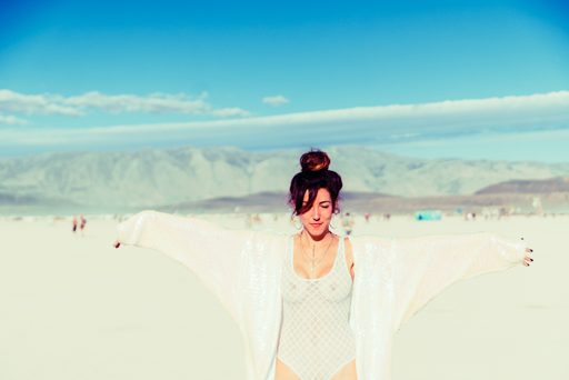 A woman with her eyes closed and arms outstretched wears a white patterned bodysuit and a shimmering white kimono-style jacket in the vast,