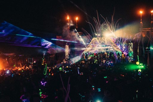 A crowd of people with glow sticks and illuminated objects enjoy a nighttime performance with lasers and pyrotechnics at the Burning Man fes