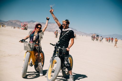 Two participants in festival attire ride fat-tire bicycles across the playa at Burning Man. The woman on the left wears a colorful beaded ne