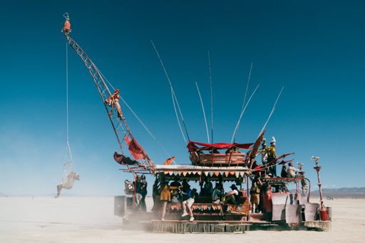 A person swings from a crane attached to a large, decorated art vehicle on the playa at Burning Man. The vehicle features multiple levels fo