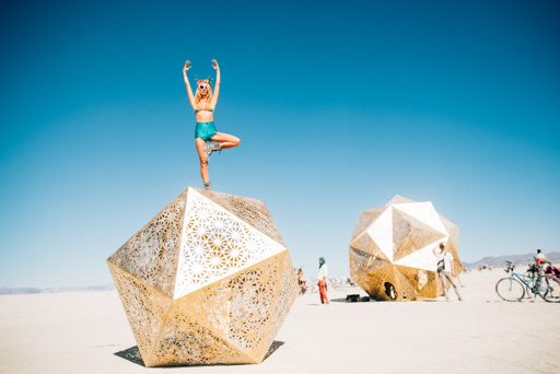 A woman performs a yoga pose atop a large, geodesic, gold-colored sculpture in the desert landscape of Black Rock City during Burning Man 20