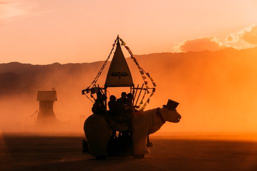 A whimsical, polar bear-shaped art car, adorned with a sail and flags, carries passengers through a dusty sunset at Burning Man 2015. In the