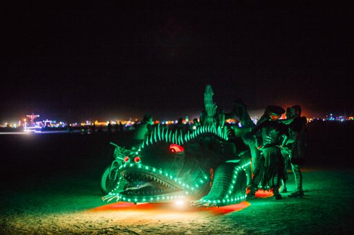 A large, illuminated dragon art car, adorned with green lights and red eyes, is parked on the playa at night during Burning Man 2015. Severa