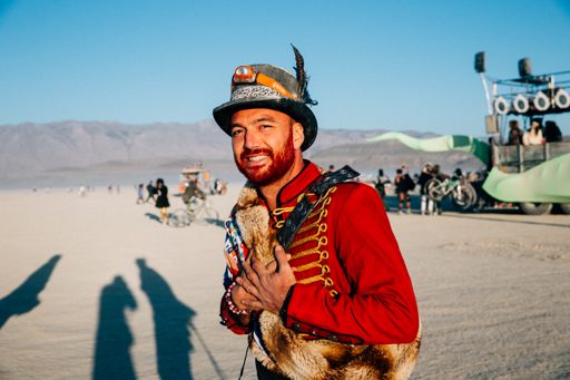 A man with red facial hair and a feathered hat poses in a red military-style jacket with gold trim and a fur vest. He is standing in a dusty