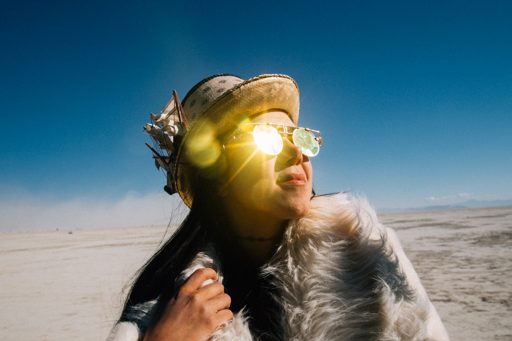 A person wearing a hat adorned with a ship's mast and sunglasses looks up at the sun, which creates a lens flare. The photograph was taken o