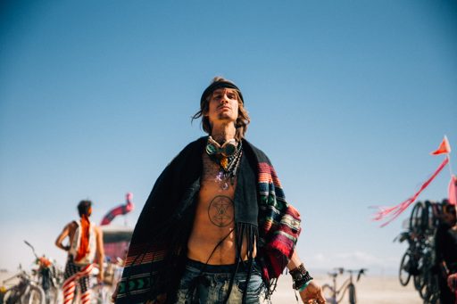A man with a star tattoo on his chest and goggles around his neck stands in the desert at Burning Man, wearing a colorful fringed shawl and