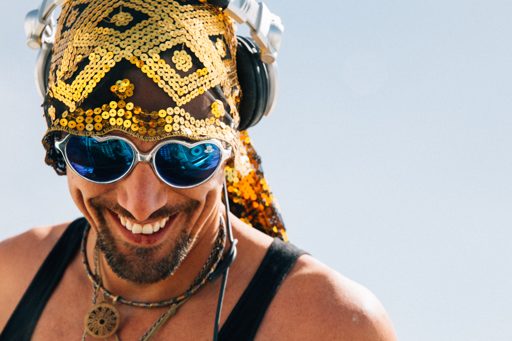 A man wearing a gold sequined bandana, heart-shaped sunglasses, and headphones smiles broadly while outdoors. The image was captured during