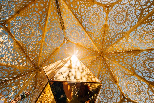 Sunlight streams through the intricate geometric golden latticework of a geodesic dome structure at Burning Man 2015, casting complex shadow