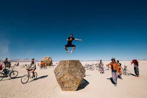 A man jumps in the air in front of a large, geometric art installation at Burning Man 2015. Other attendees on bicycles and in the backgroun