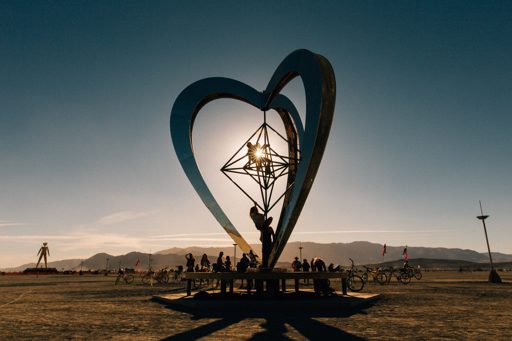 The sun sets behind the "Heart of the City" sculpture at Burning Man, casting long shadows across the playa. Silhouettes of attendees and bi