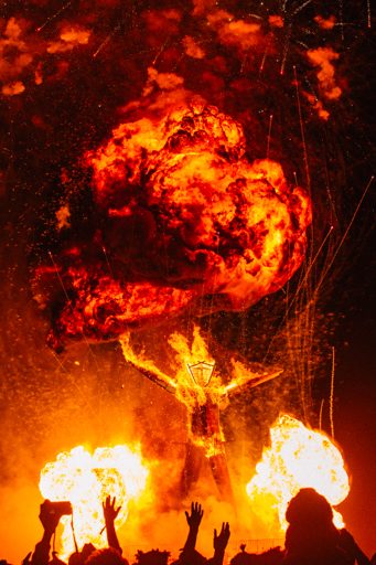 A large effigy, engulfed in flames, is raised by a crowd at the Burning Man festival. Explosions and fireworks erupt around the central figu