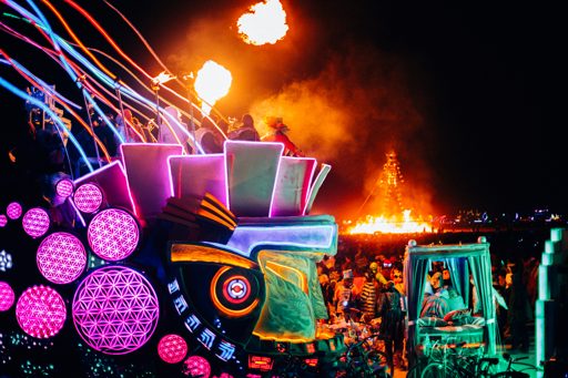 A vibrant night scene at Burning Man 2015 features illuminated art installations with geometric patterns and colorful lights, set against a