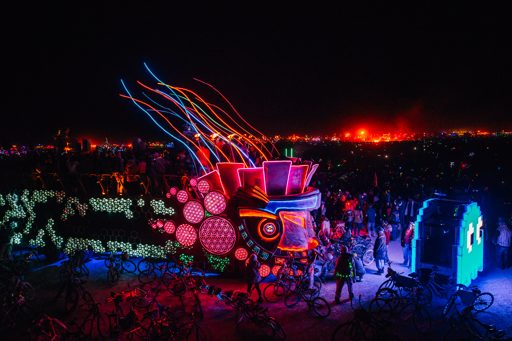 A nighttime scene at Burning Man 2015 features illuminated art installations, including a large, colorful structure with neon lights and a p