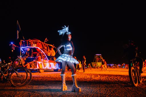 A woman in a light-up costume poses in front of a decorated art car at night during Burning Man 2015. The scene is illuminated by numerous c