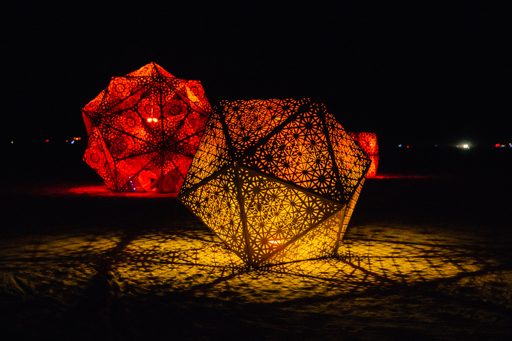 Two large, illuminated geodesic dome sculptures cast intricate shadows on the dark ground at the 2015 Burning Man festival. The sculpture in