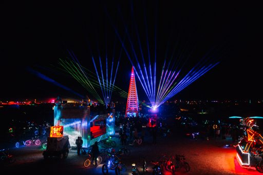 Laser light beams project from a tall wooden structure and illuminate the night sky over a crowd of people and illuminated art installations