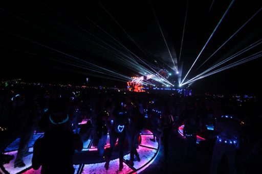 A crowd of people dance under a canopy of laser lights at night in Lovelock, Nevada. Many attendees wear glowing accessories, and the ground