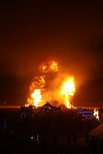 A large structure erupts in flames and smoke at night during the Burning Man festival in Lovelock, Nevada. A crowd of spectators watches the
