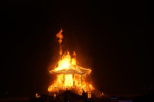 A hexagonal pavilion engulfed in flames at night during the Burning Man festival in Lovelock, Nevada. The structure is illuminated by the in