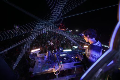 A DJ performs at night on a stage illuminated by numerous laser beams, with a large crowd visible in the background. The DJ is operating a m