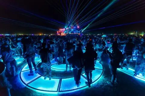 A crowd of people dance under a canopy of blue and red laser lights at night. They are gathered on a circular platform illuminated with blue