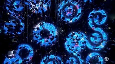 An aerial view captures a crowd of people walking on illuminated circular platforms at night. The platforms are lit with bright blue neon li