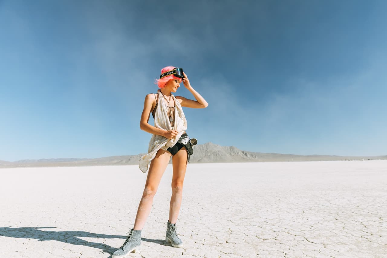 A woman with vibrant pink hair and steampunk-inspired attire stands in a vast, cracked desert landscape under a clear blue sky.