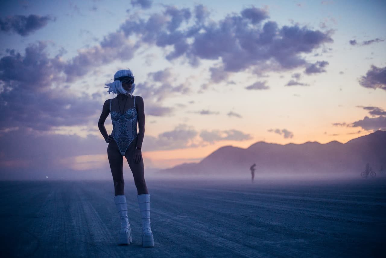 A person in a white futuristic costume stands dramatically on a vast, dusty desert playa at sunset, with mountains in the distance.