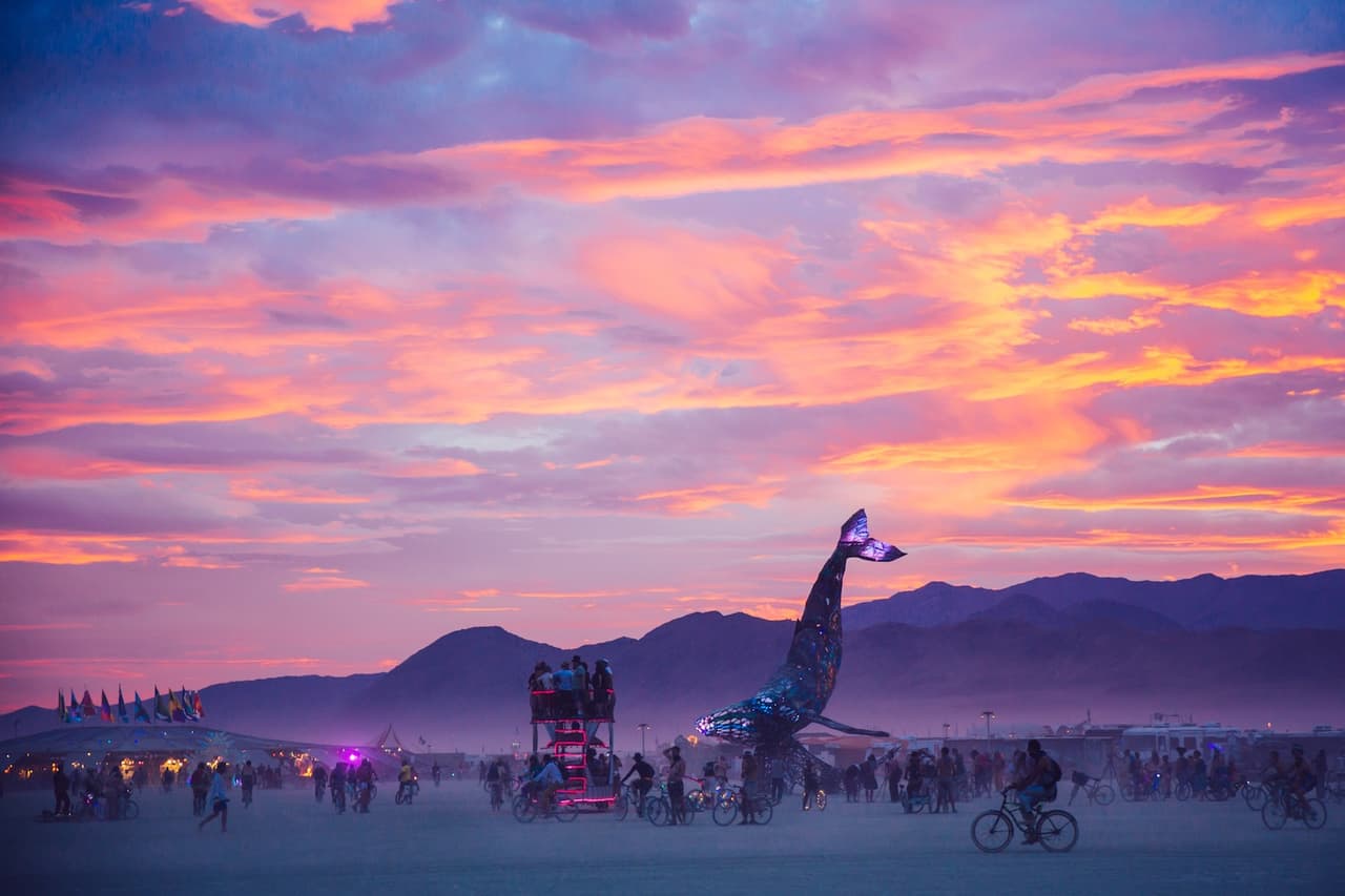 A vibrant sunset sky with hues of orange, pink, and purple illuminates a large, glowing whale sculpture surrounded by people and bicycles in