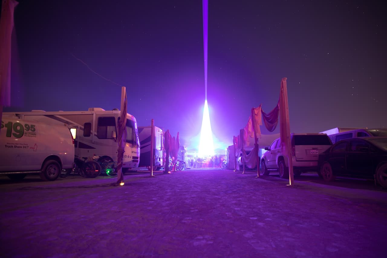 A powerful purple laser beam shoots into the dark night sky from a distant structure, flanked by parked RVs and vehicles on a wide, dusty pa