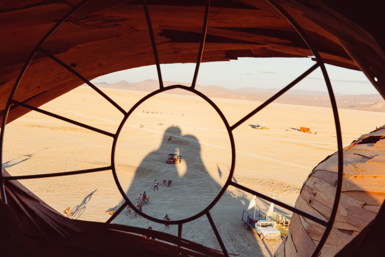Two kissing shadows are cast onto a vast desert landscape, viewed through a circular, spoked window frame.