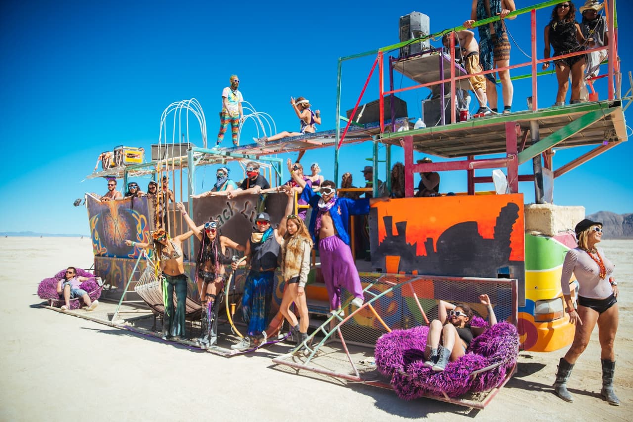 A large group of people in eclectic attire pose on and around a multi-level art car in a vast, sunny desert landscape.