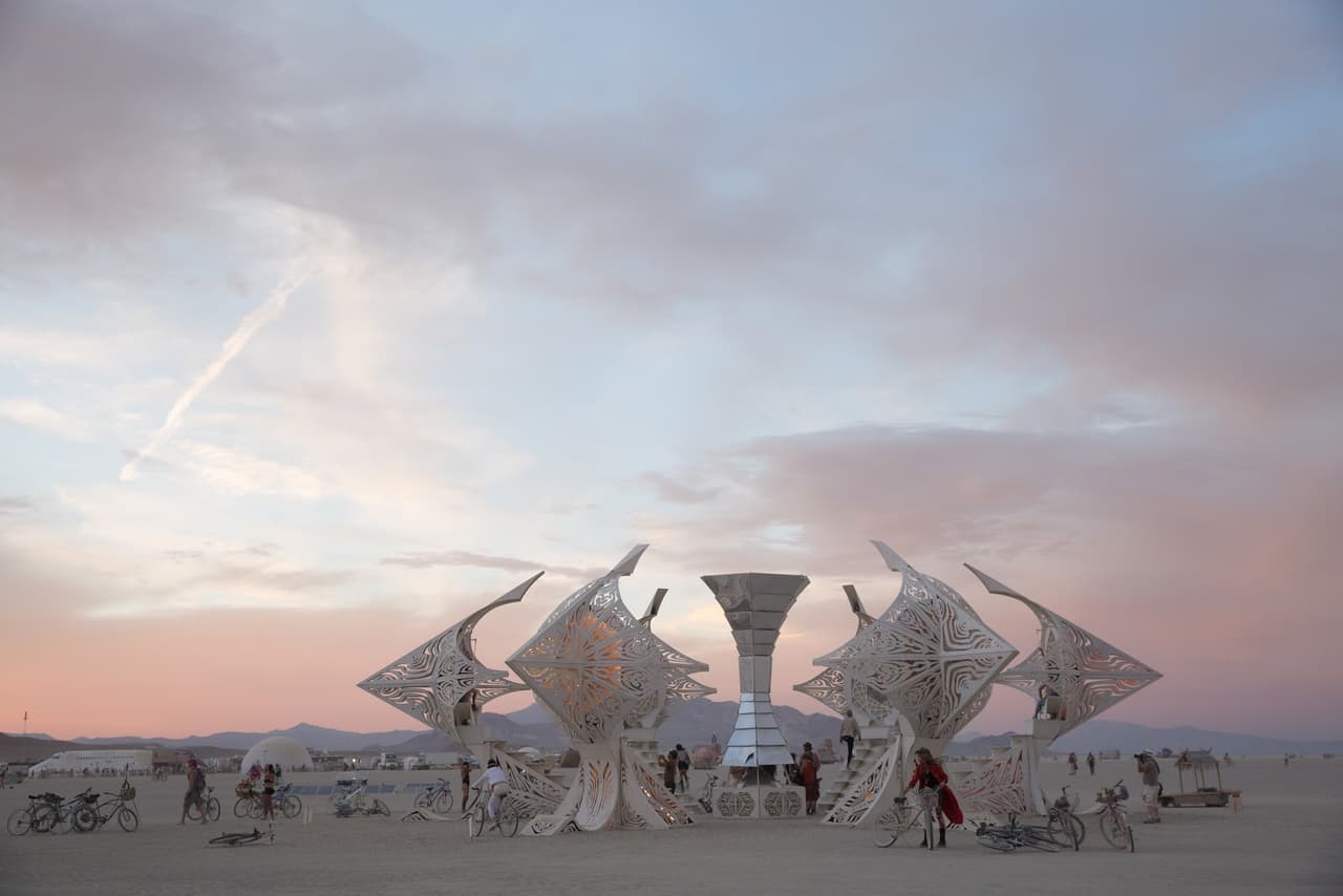 A large, intricate white geometric art installation stands prominently in a vast desert landscape under a soft, colorful sunset sky.