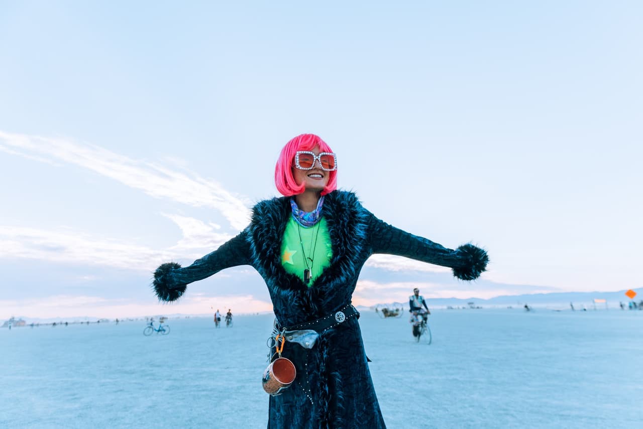 A person with vibrant pink hair and a furry coat stands with arms outstretched on a vast, pale playa under a bright sky.