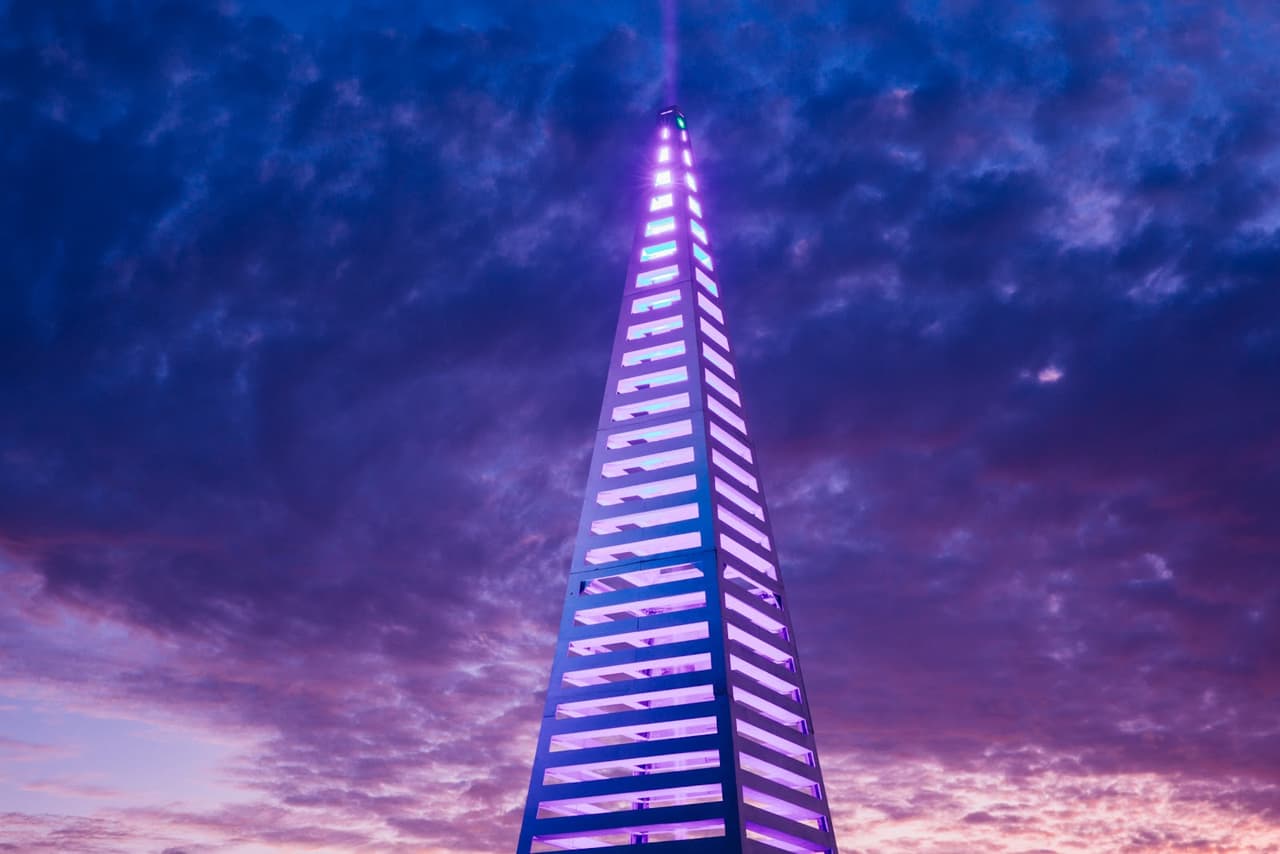 A tall, slender skyscraper glows with vibrant purple light against a dramatic twilight sky.