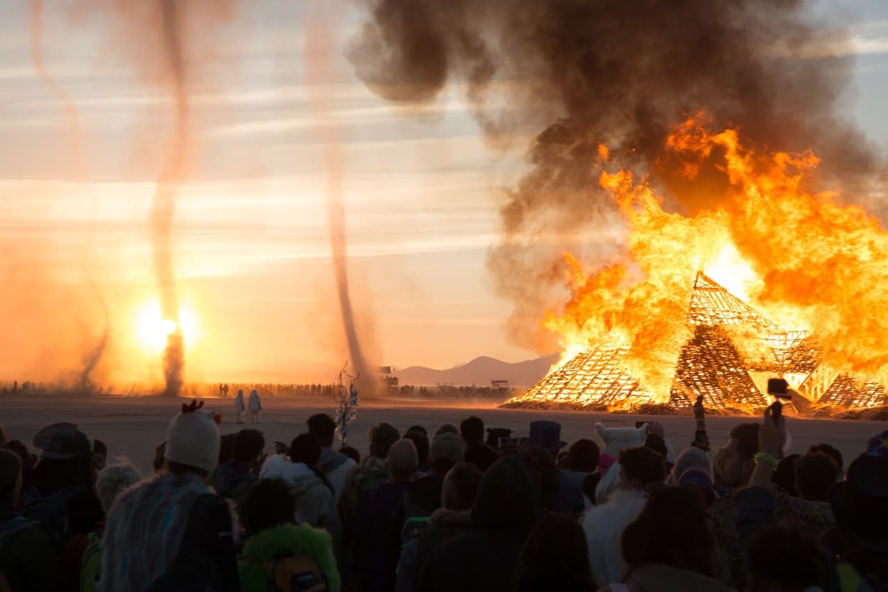 A massive wooden structure burns fiercely at sunset, while two large dust devils rise into the sky over a crowd of spectators.