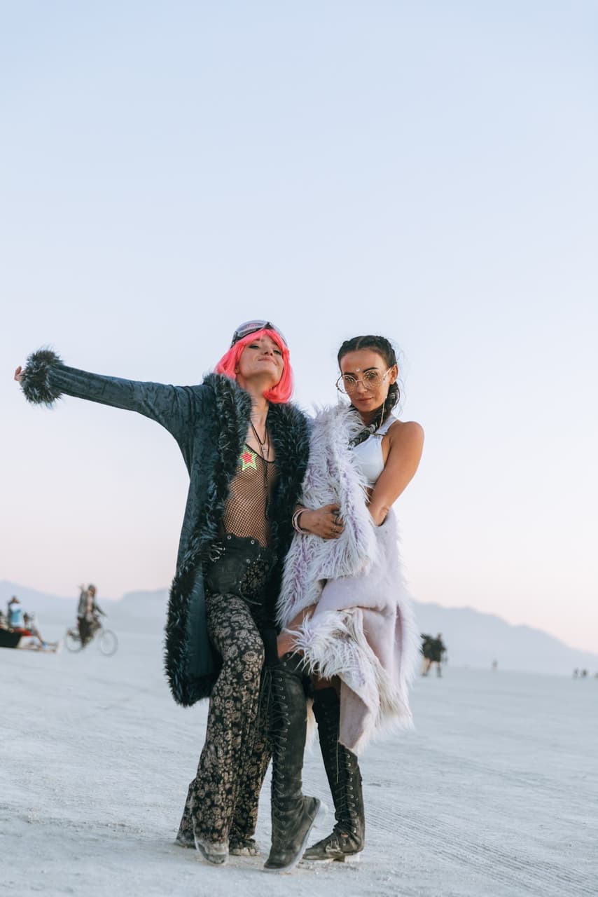 Two women in elaborate, festival-style outfits pose on a vast, flat desert landscape under a pale sky.