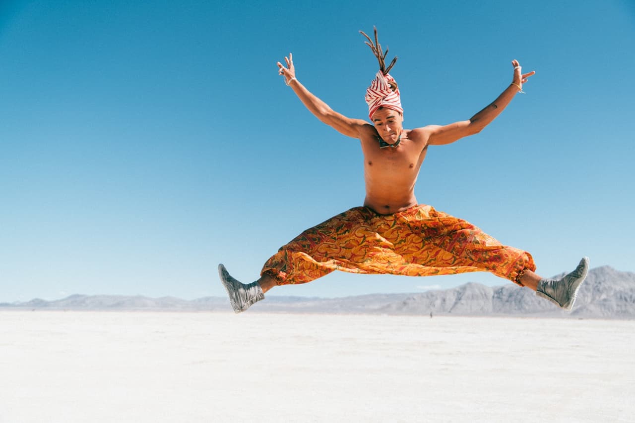 A person in vibrant orange pants and a unique headwrap leaps joyfully against a vast desert and clear blue sky.