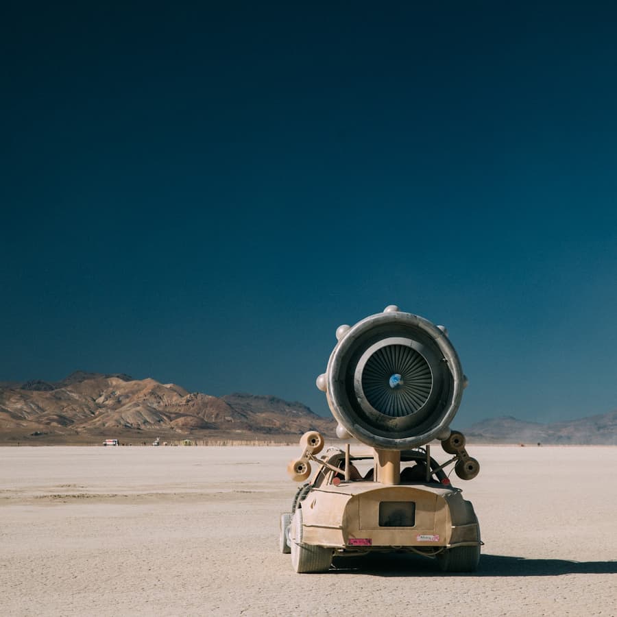 A unique, custom-built desert vehicle with a large jet engine mounted on its rear drives across a vast, flat playa under a clear blue sky.