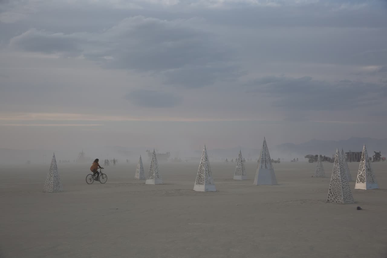 A lone person cycles through a hazy desert landscape dotted with white, pyramid-like art installations under an overcast sky.