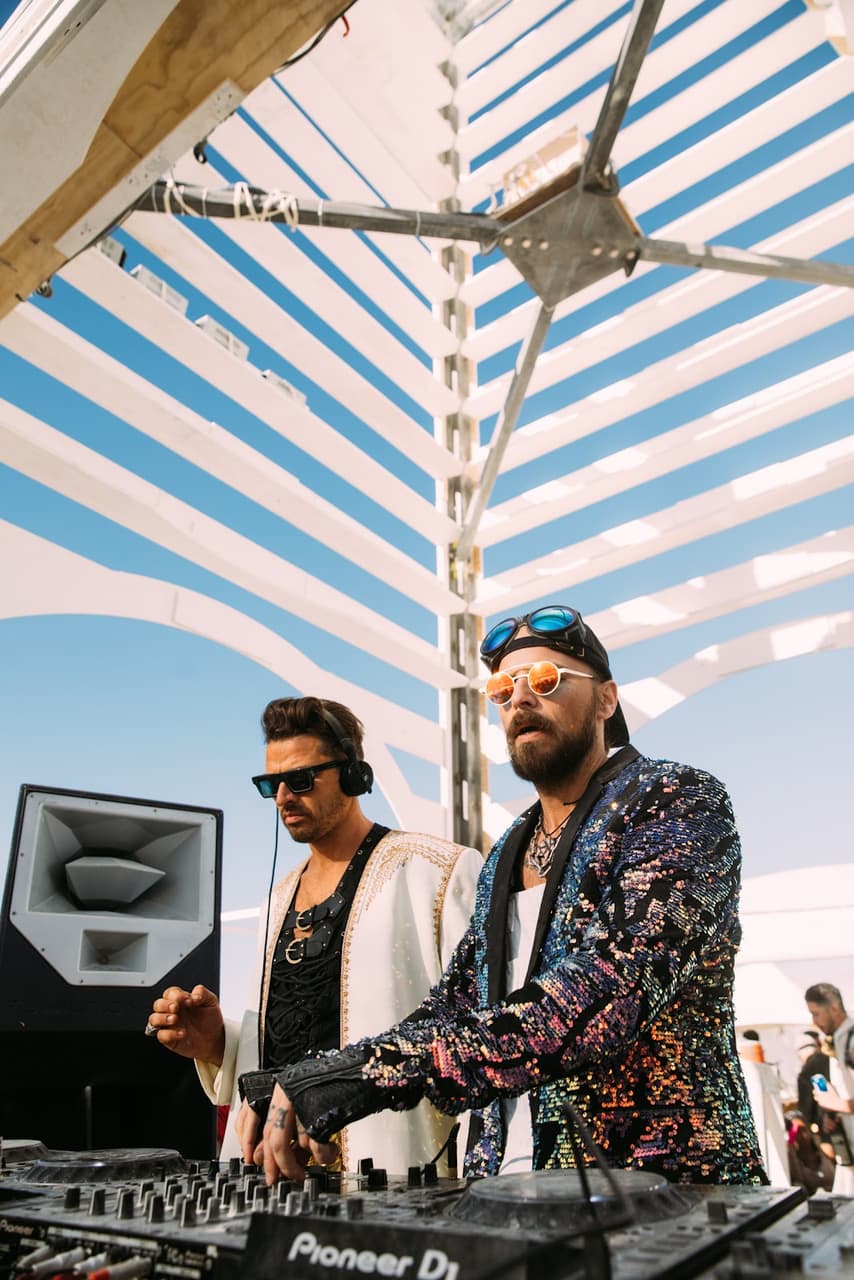 Two stylish DJs perform an outdoor set under a striped canopy, with one wearing a sequined jacket and the other a white robe.